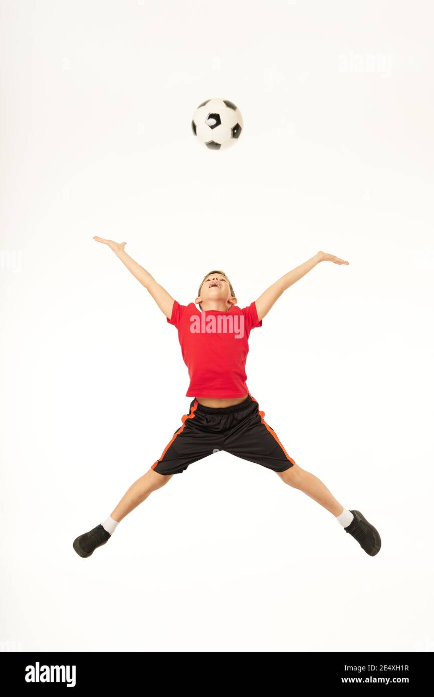 Cute boy football player throwing up game ball and smiling while jumping in the air. Isolated on white background Stock Photo