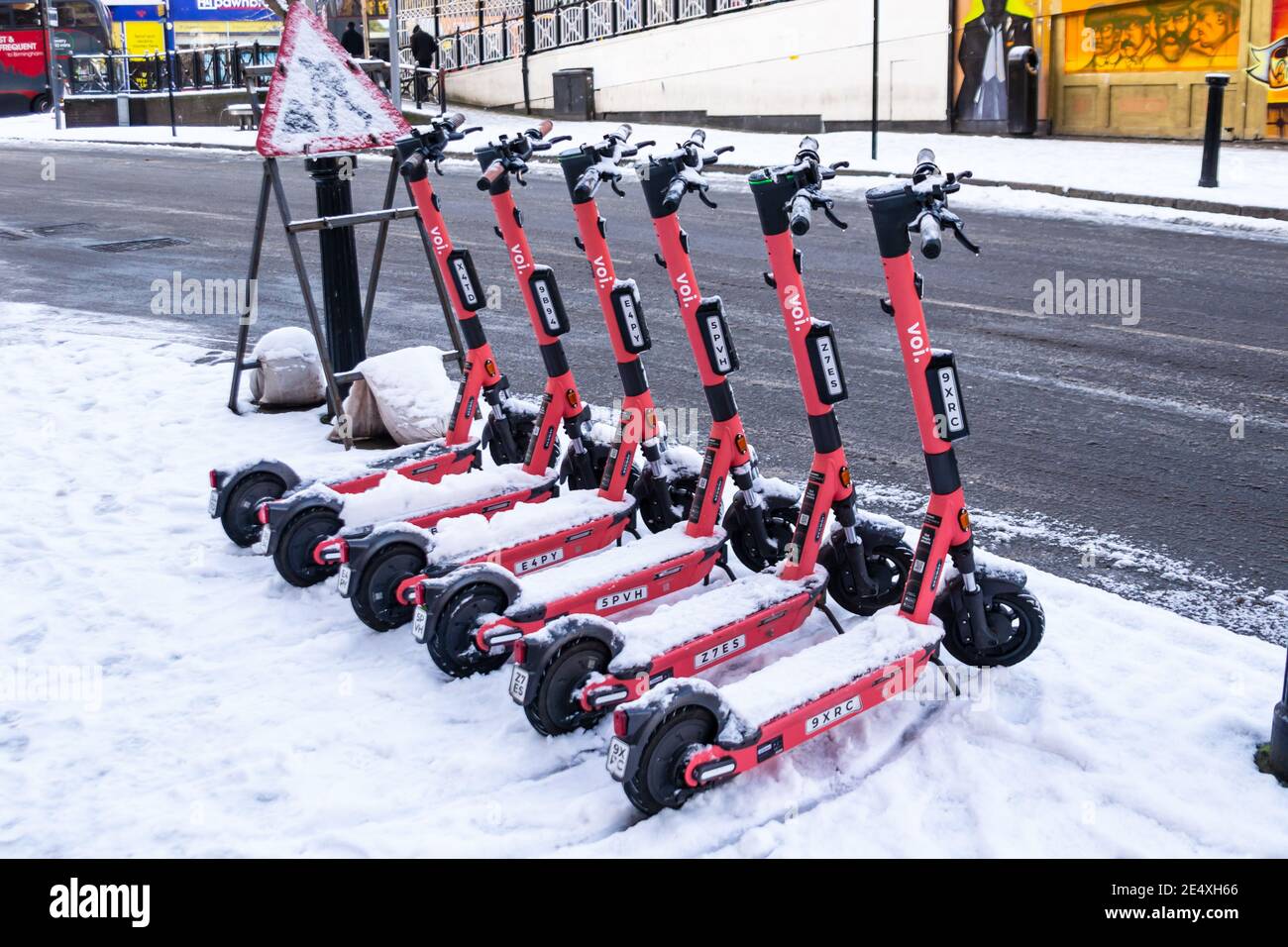Birmingham, West Midlands, UK. 25th January 2021 - Voi electric scooters lined up in the snow on Dale End across the street from the Peaky Blinder pub. Credit: Ryan Underwood / Alamy Live News Stock Photo