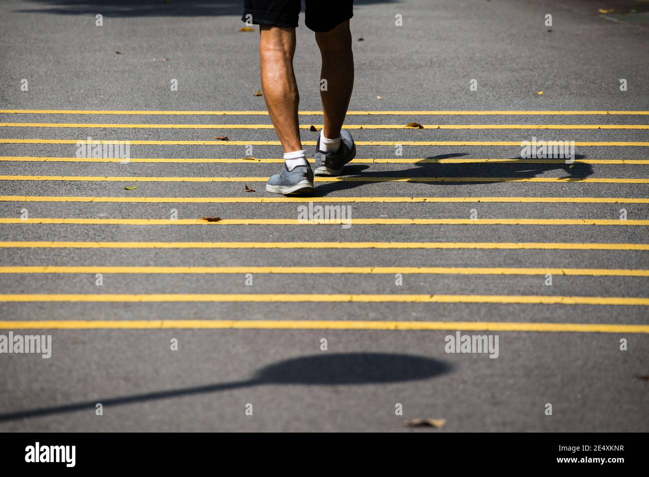 A pair of aging strong legs walking on yellow strips tarmac pavement and his contrasty body shadow on the ground. Singapore Stock Photo