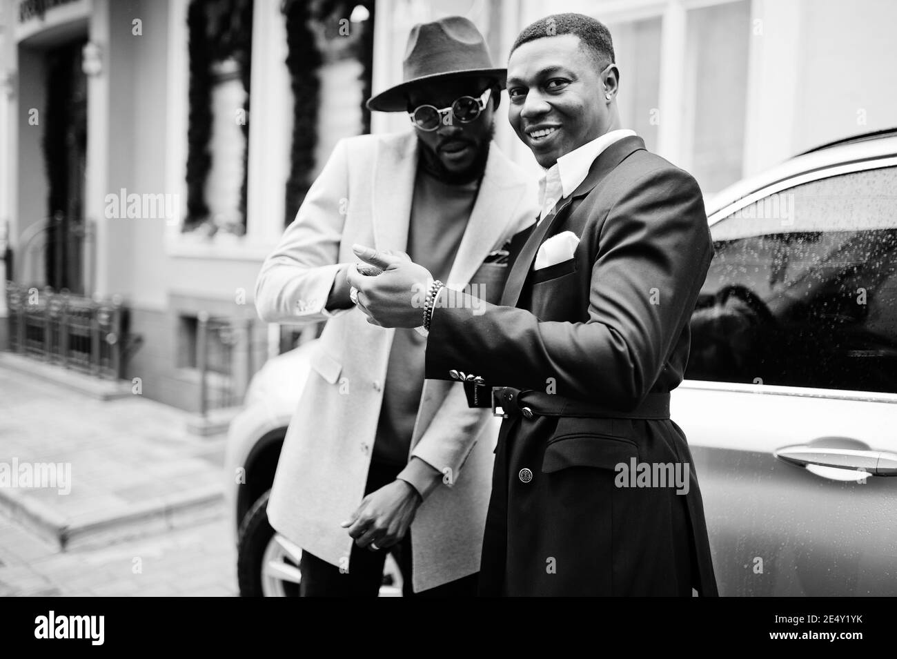 Two fashion black men stand near business car and look at cell phone. Fashionable portrait of african american male models. Wear suit, coat and hat. Stock Photo