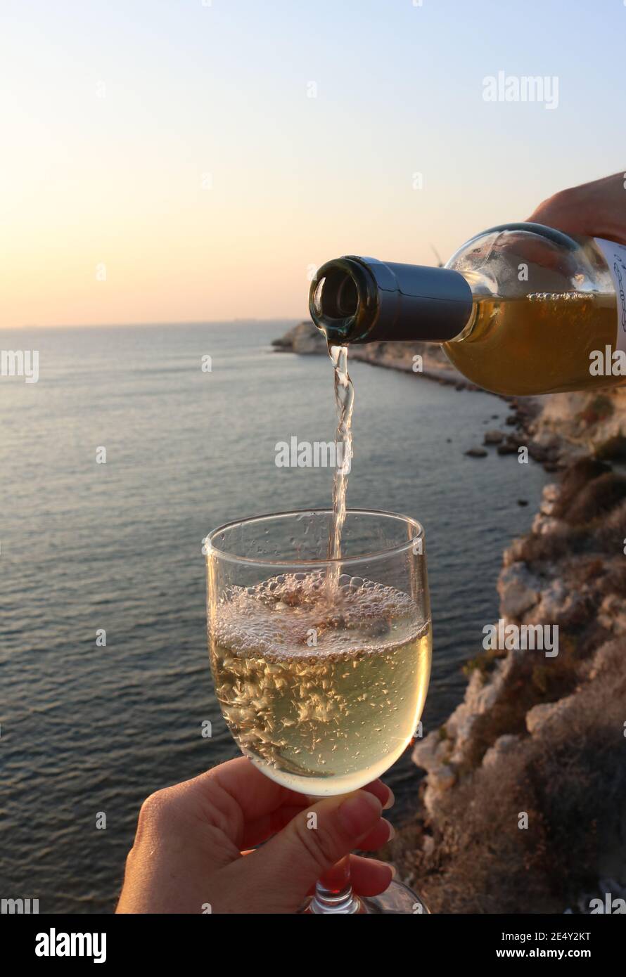 Close up of a man pouring white wine in a glas with a beautiful scenery of Bozcaada island at sunset in the background Stock Photo