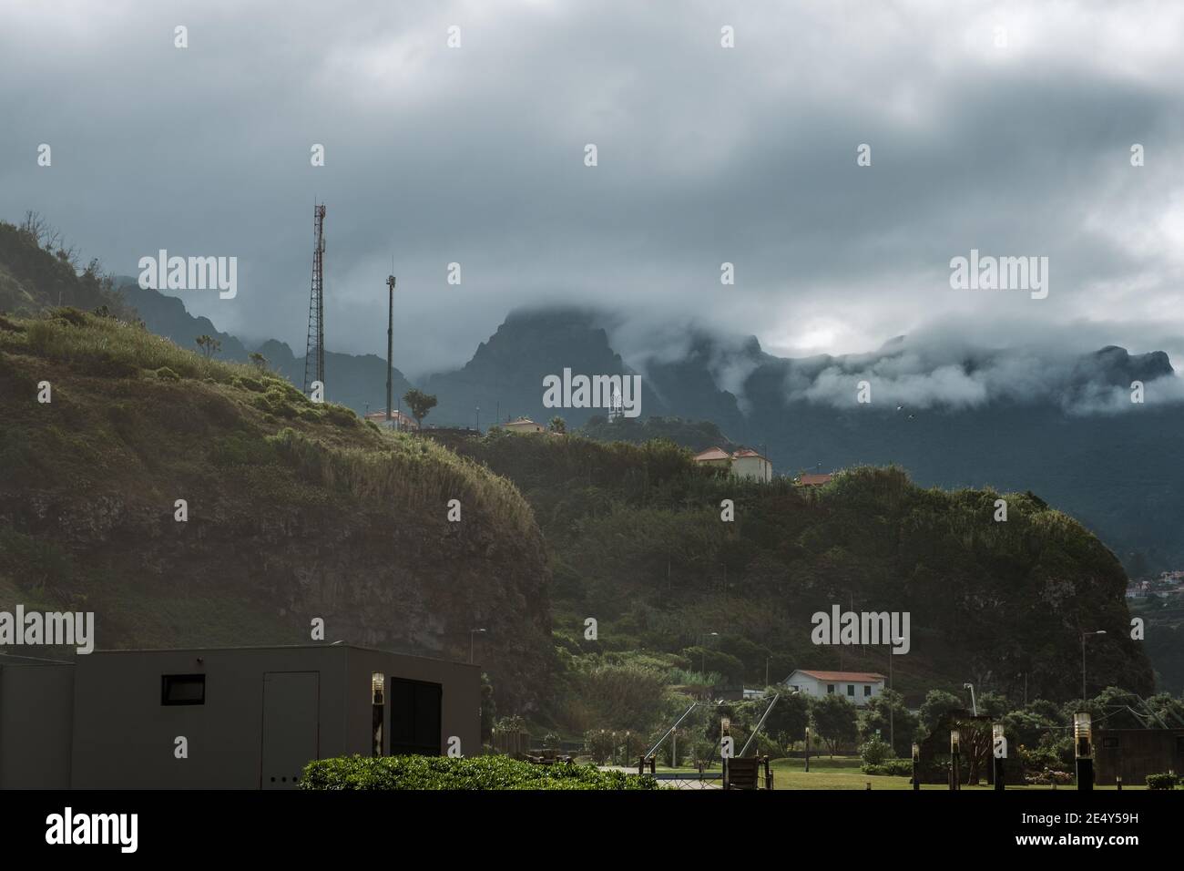 small churche and houses at top of a hill wth a mountain range on the background covered by clouds Stock Photo