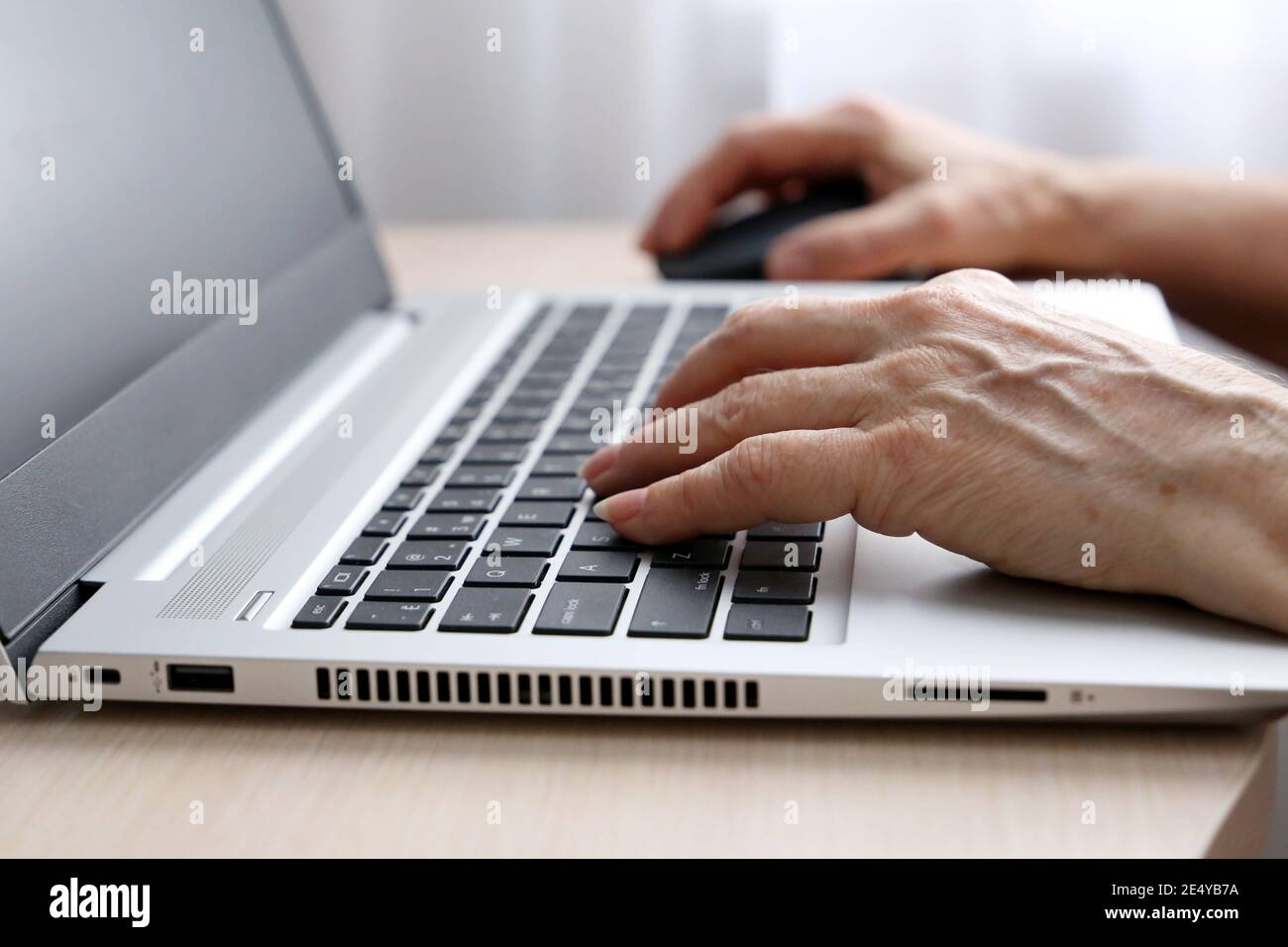 Elderly woman working at the laptop. Wrinkled female hands on notebook keyboard on a desk in sunlight Stock Photo