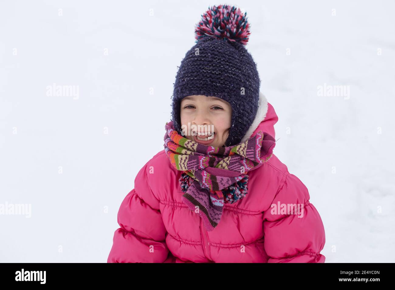 A cute little girl in a pink jacket and a hat is playing in the snow ...