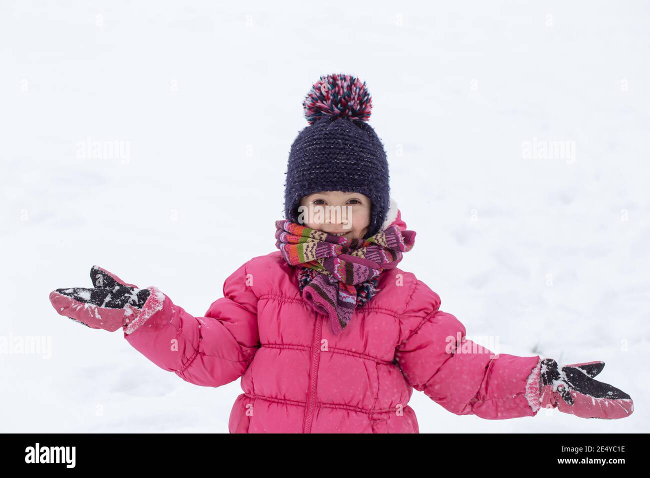 A cute little girl in a pink jacket and a hat is playing in the snow ...