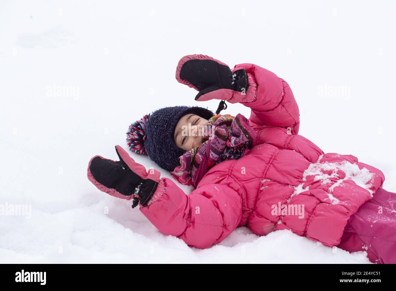 A cute little girl in a pink jacket and a hat is playing in the snow ...