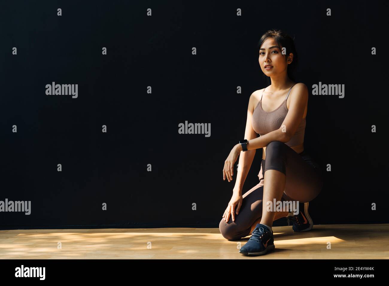 Focused young Asian sportswoman relaxing and getting ready for workout while crouching in studio on black background - full body length Stock Photo