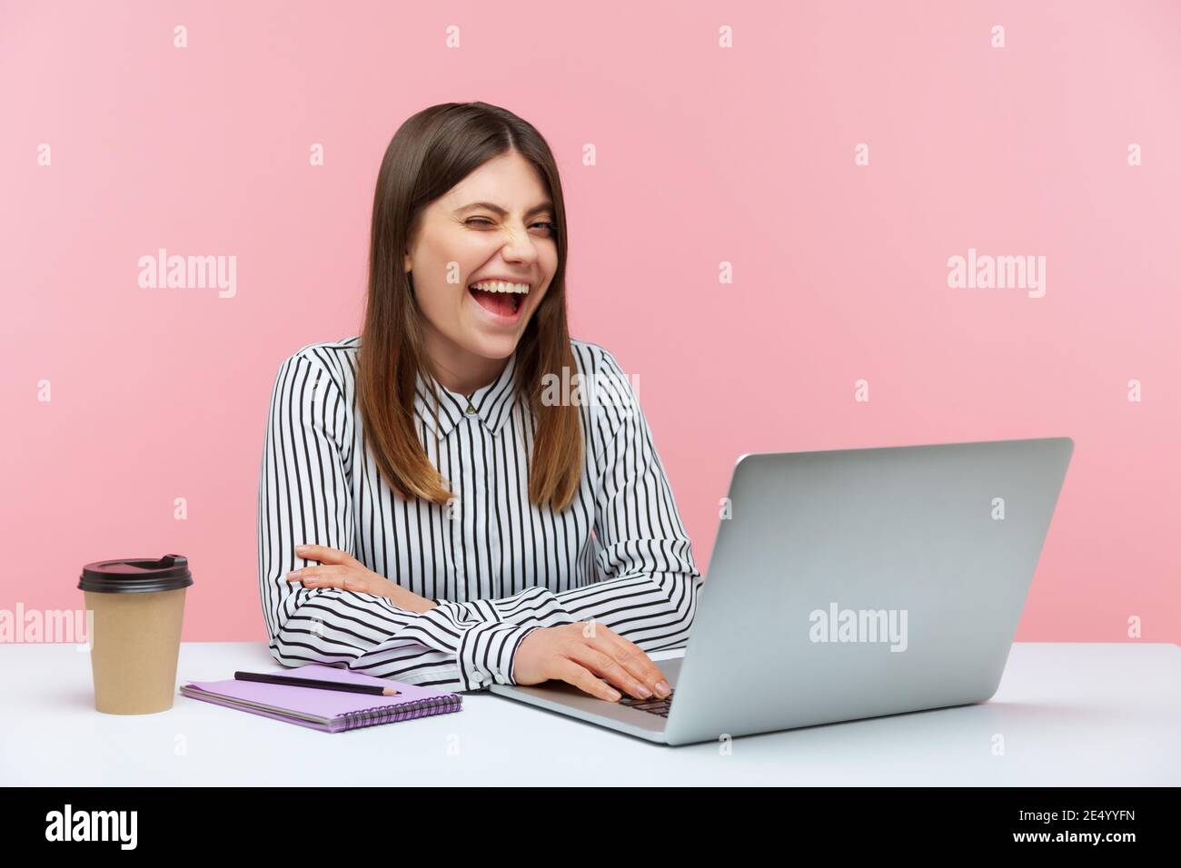 Mischievous excited woman office worker in striped shirt sitting at workplace and winking, looking at camera with toothy smile, good mood, happiness. Stock Photo