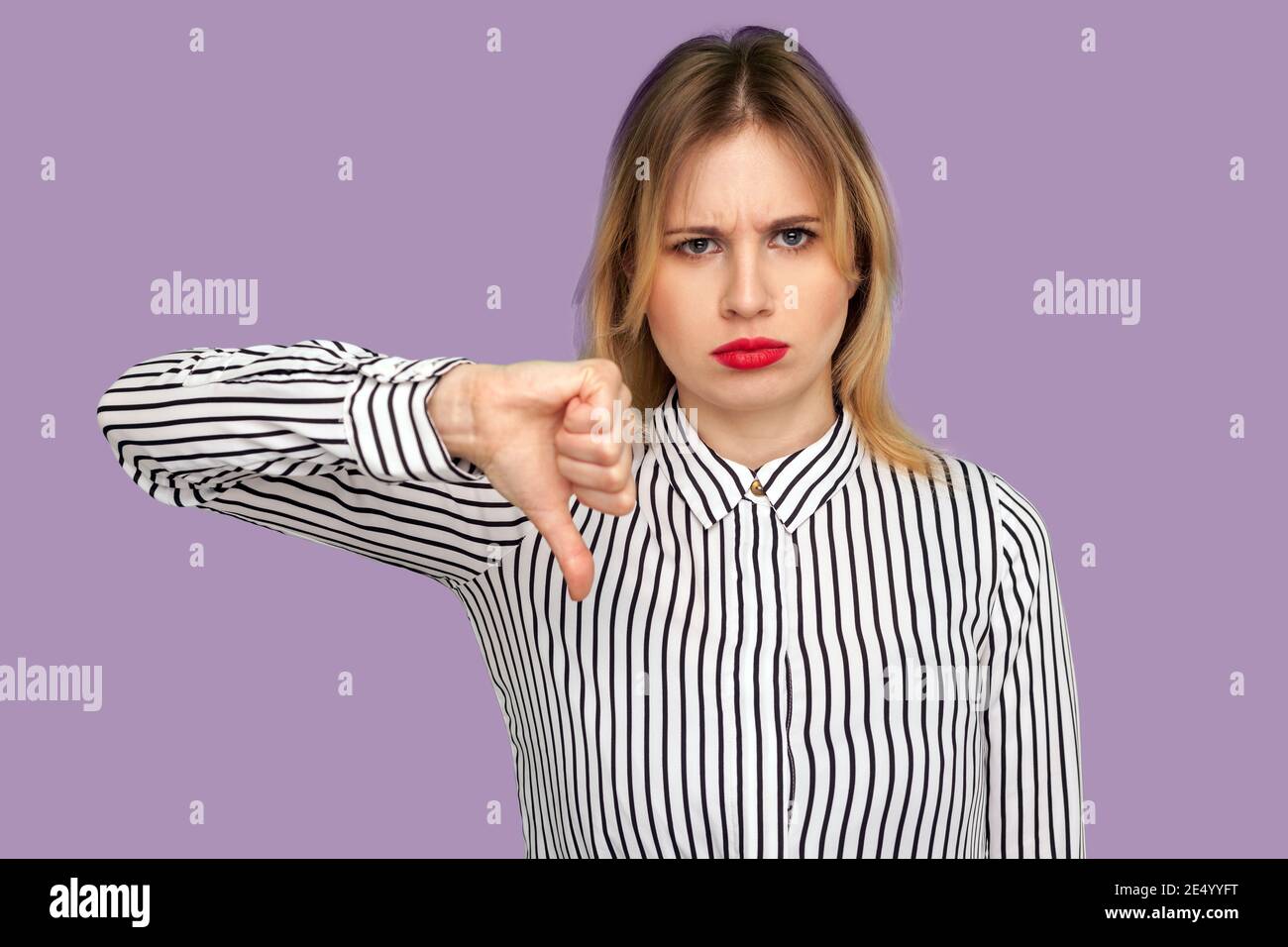 Dislike! Attractive blond woman frowning with naughty expression and showing thumbs down, expressing disapproval, negative feedback. indoor studio sho Stock Photo