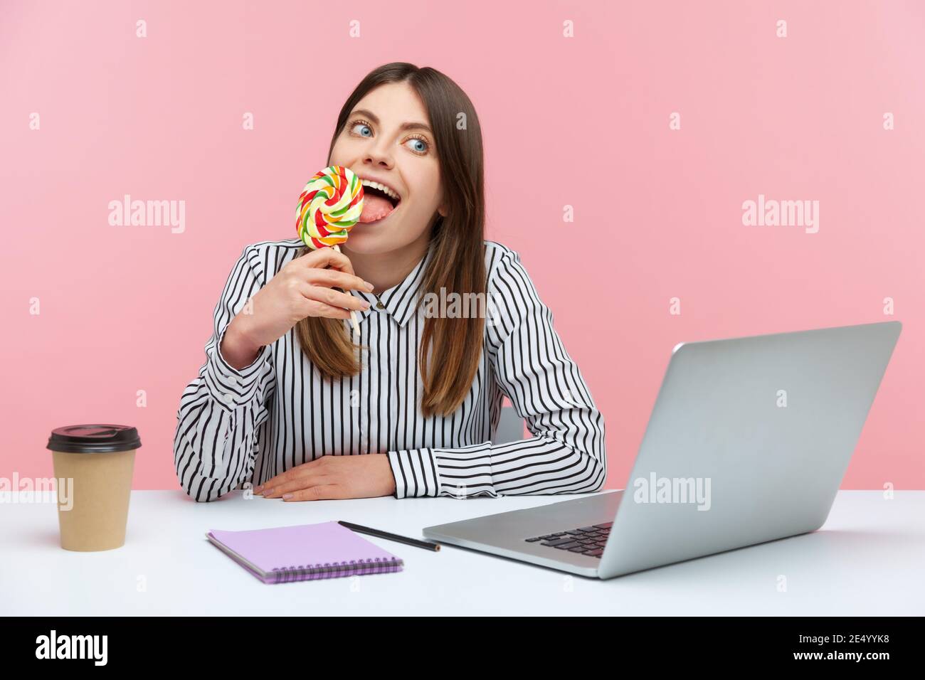 Dreamy funny woman office worker licking big striped lollipop sitting at workplace with laptop, enjoying sweets during break. Indoor studio shot isola Stock Photo
