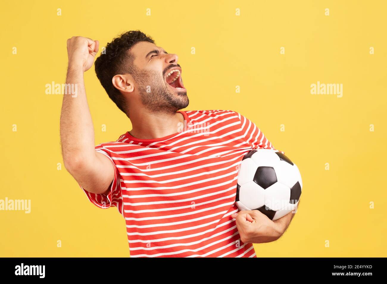 Extremely positive satisfied man in striped red t-shirt screaming holding soccer ball, celebrating victory of favourite football team on championship. Stock Photo