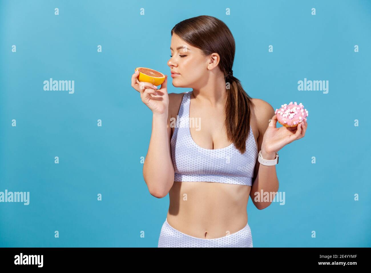 Sportive woman in white top holding in hands half of juicy grapefruit and round donut with pink icing, enjoying fresh aroma of ripe fruit. Indoor stud Stock Photo