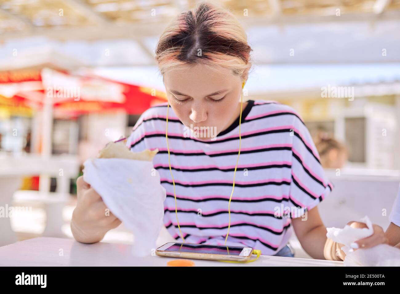 Teen girl eating a sandwich, burger, looking at the smartphone screen in headphones Stock Photo