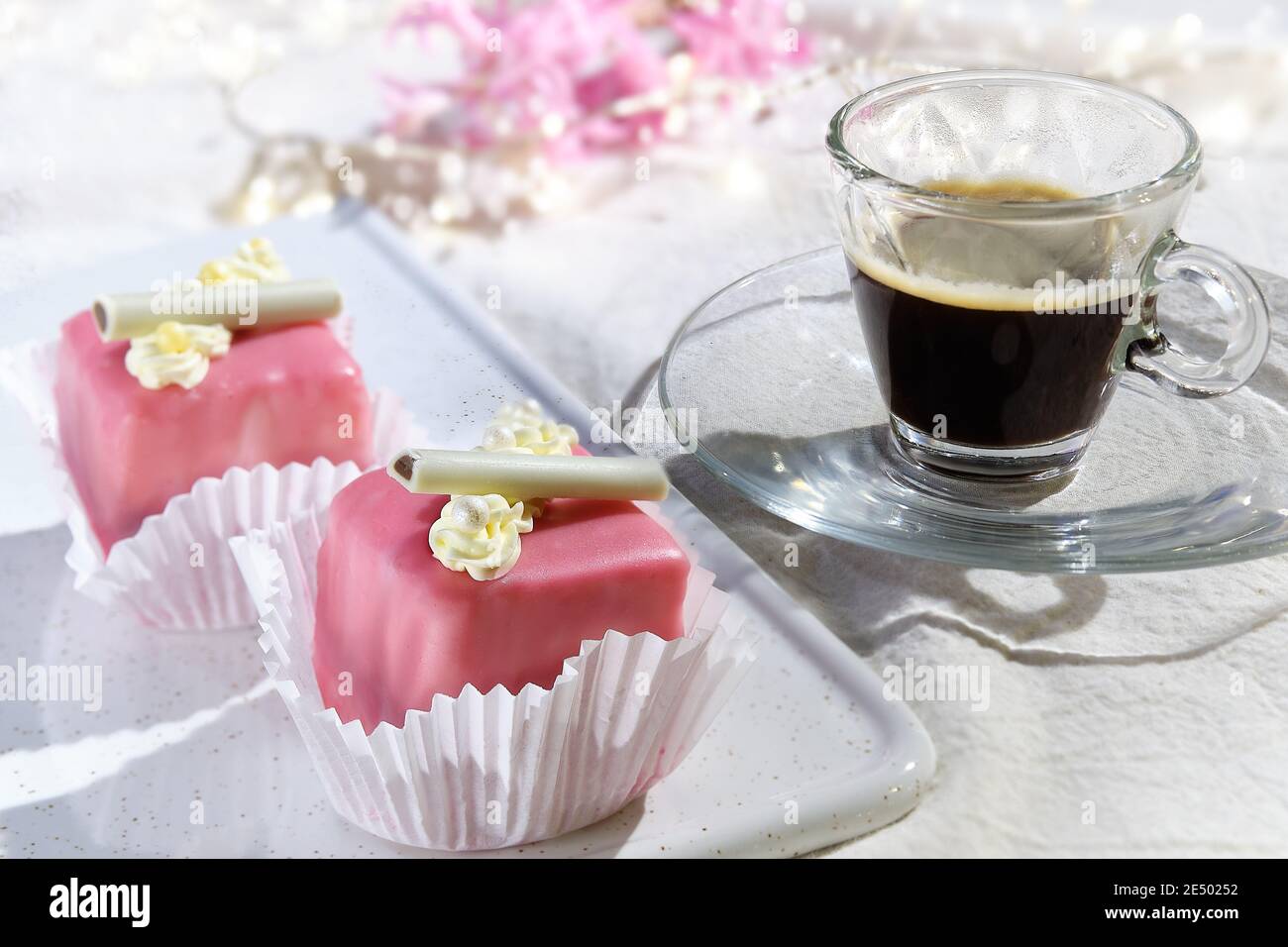 Valentine petit fours with marzipan icing and cream flowers. Espresso coffee in glass cup. Garland of lights on white textile. Pink hyacinth flower Stock Photo