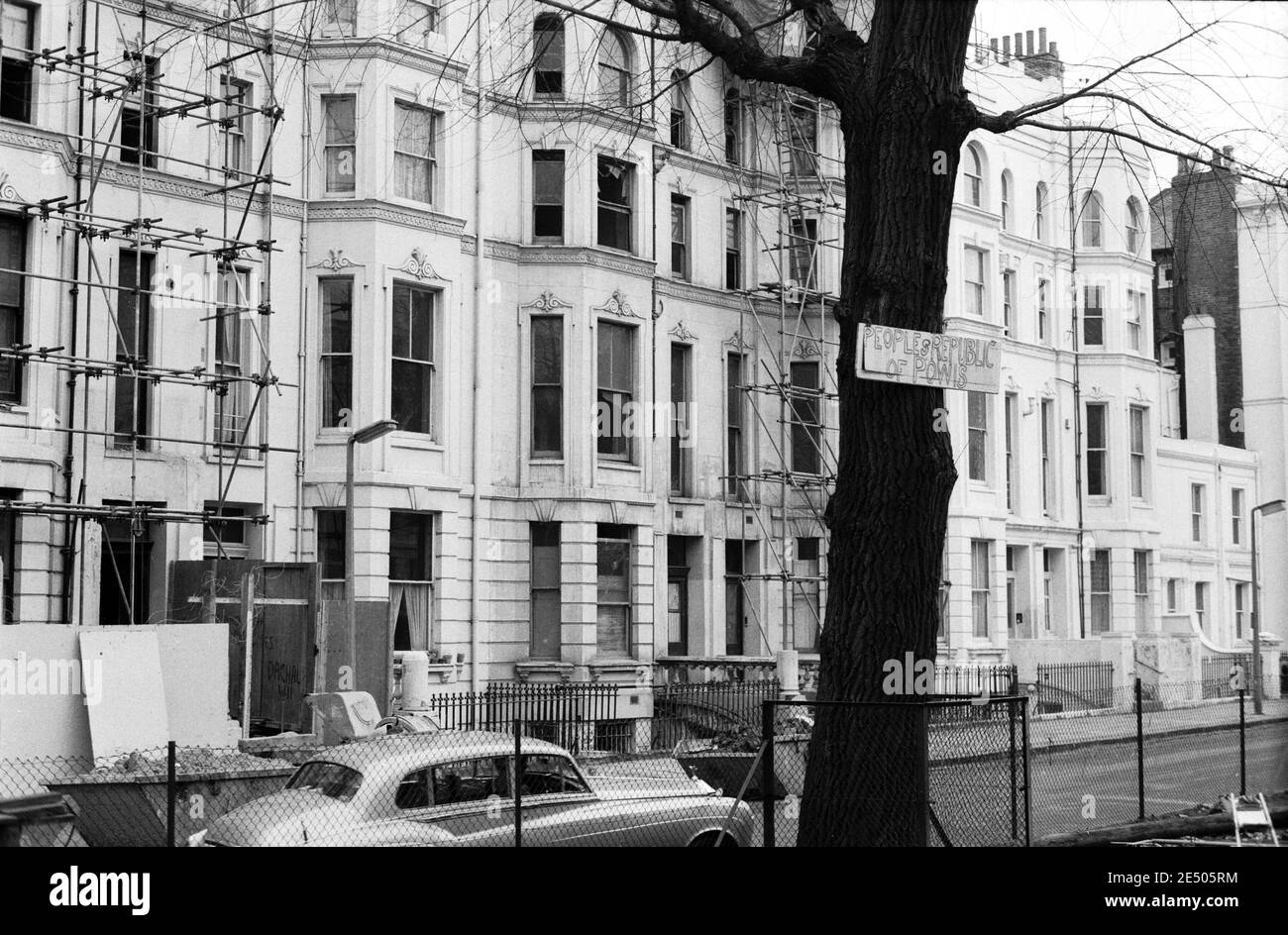 UK, West London, Notting Hill, 1973. Rundown & dilapidated large four-story houses are starting to be restored and redecorated. Facing approximately No.20 Powis Square from Powis Square play area. The sign on the tree reads 'Peoples Republic of Powis'. A Rolls Royce car is parked opposite scaffolding. Stock Photo