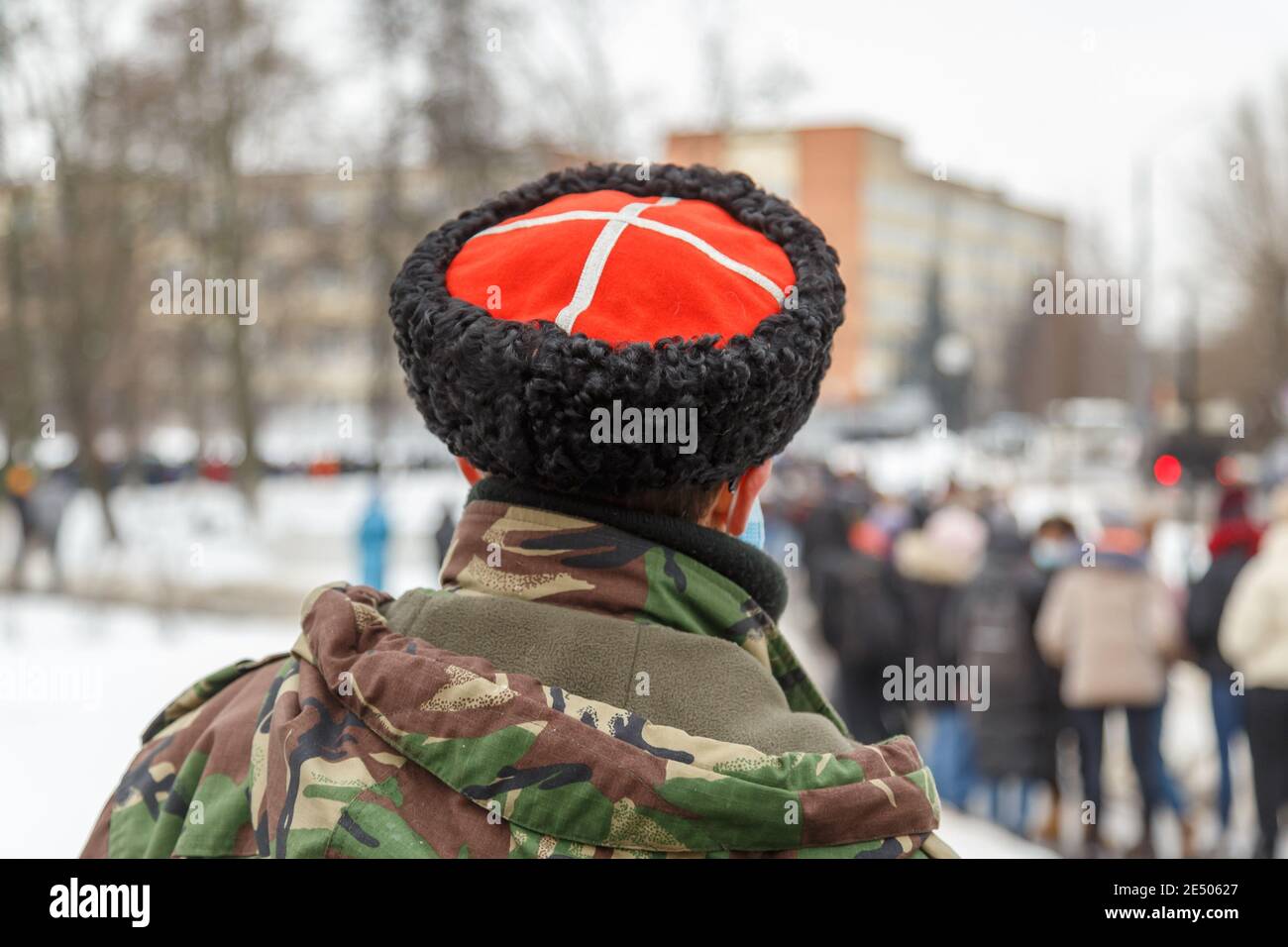 man in camouflage jacket and cossack hat with white cross on red watching blurry crowd of people - view from back. Stock Photo