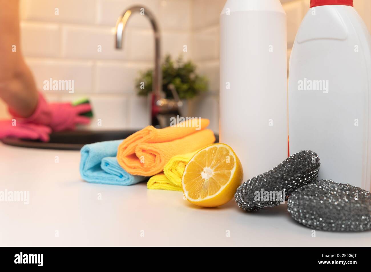 Cleaning products, citron and sponges on kitchen countertop with in focus and in background blurred womens hands cleaning kitchen. Stock Photo