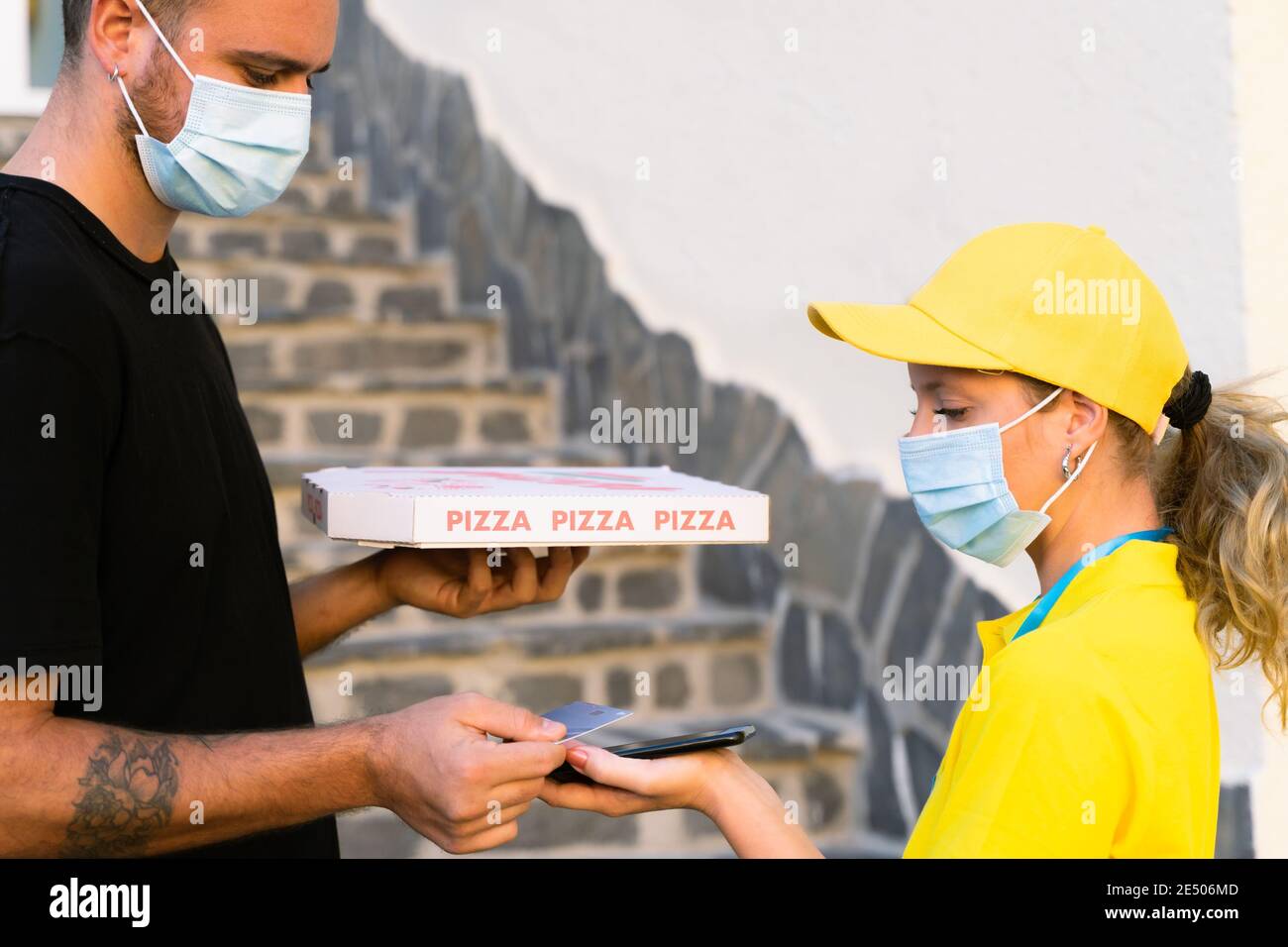 Delivery woman employee in yellow cap and t-shirt uniform face protective mask. Man hold a pizza box and pay with credit card. Coronavirus and buy con Stock Photo
