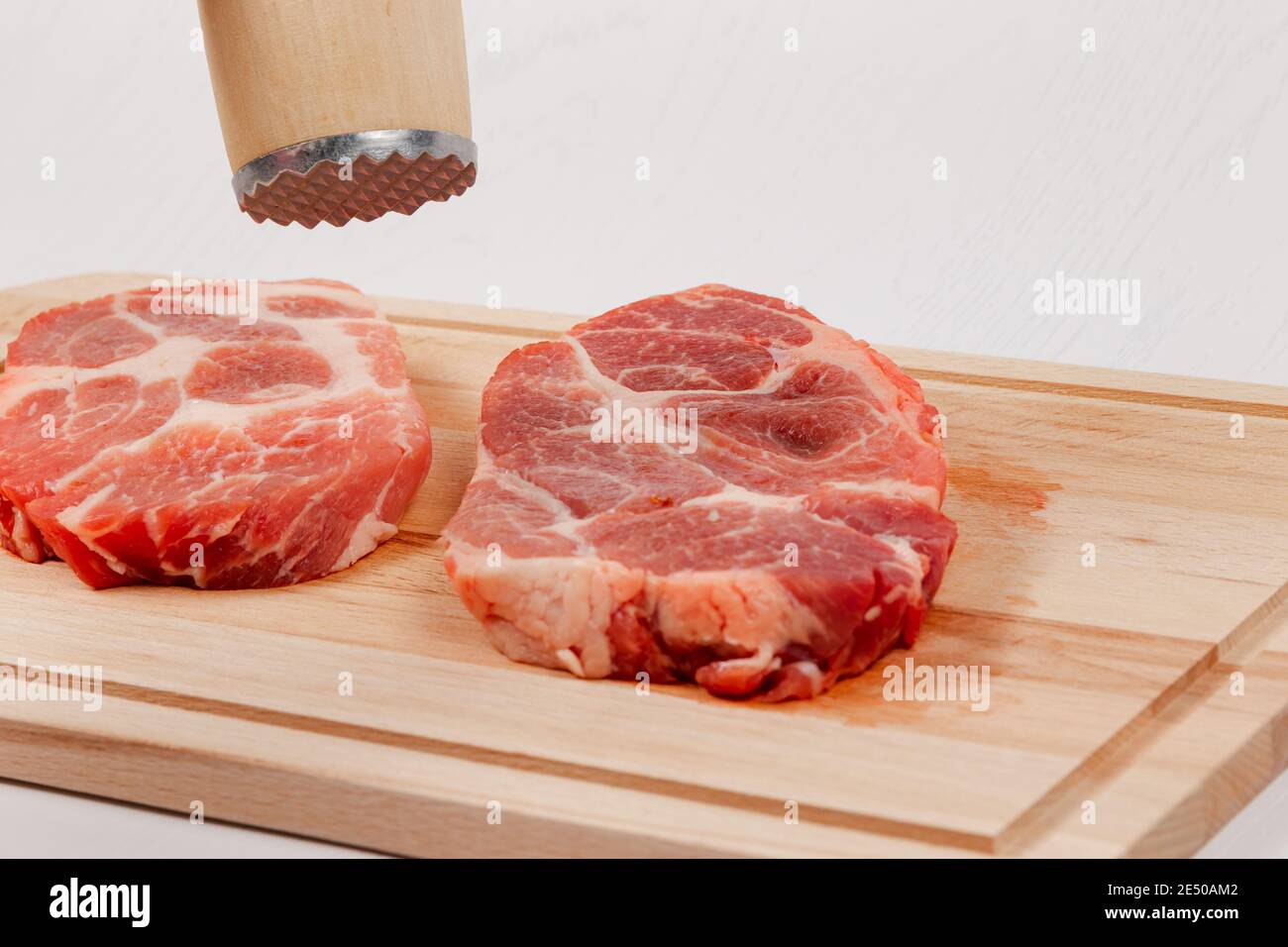 https://c8.alamy.com/comp/2E50AM2/fresh-sliced-raw-pork-steaks-meat-on-a-cutting-kitchen-board-on-a-white-wooden-table-above-them-a-hammer-for-beating-meat-2E50AM2.jpg