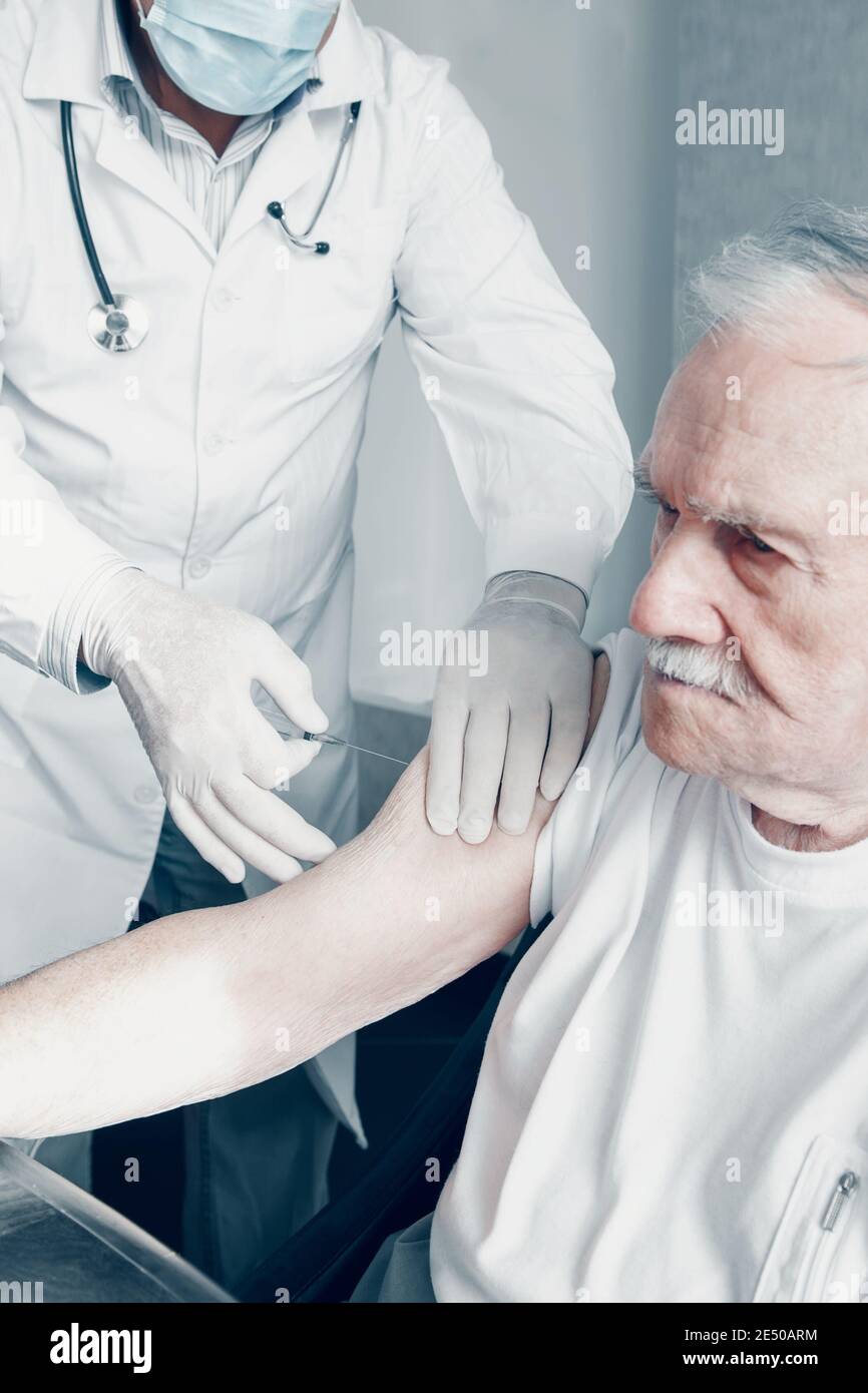 Vaccination of the elderly against viruses. A medical worker gives an injection of the covid-19 vaccine to an old, white-haired man. The old man looks Stock Photo