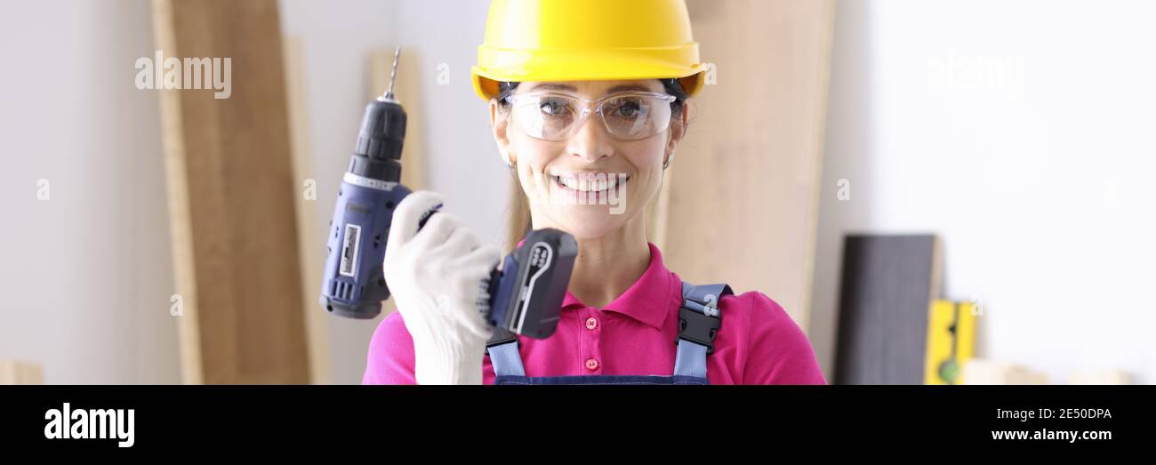 Woman builder in overalls and protective helmet holding drill in hands in workshop Stock Photo
