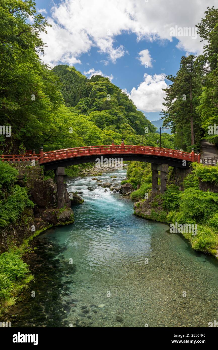 Wide angle view of a japanese landscape with green sloping hills, a red wooden bridge over a brook, against a blue sky with clouds Stock Photo