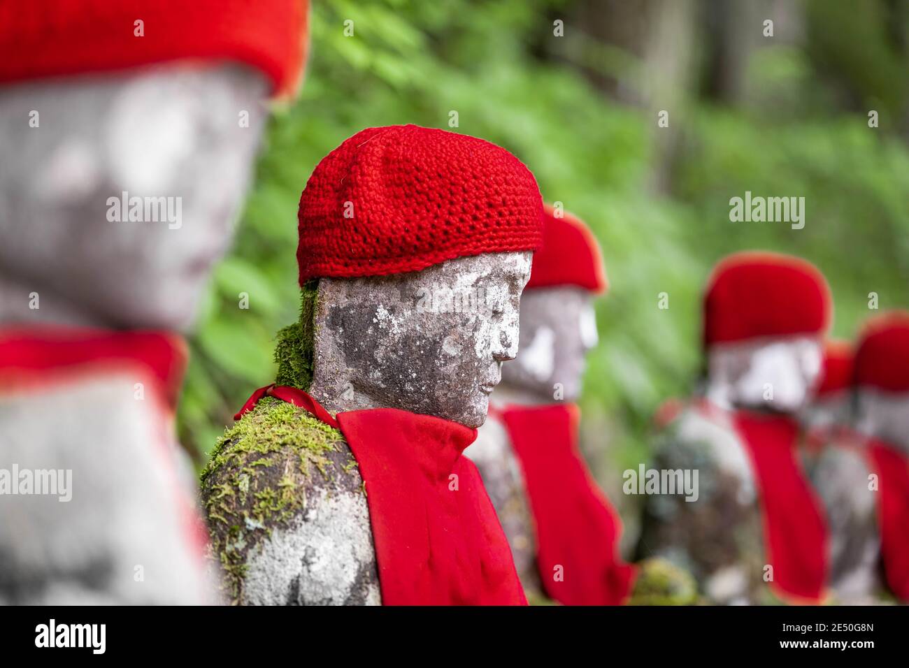 Close up of a row of stone japanese jizo statues wearing red caps and bibs, against a green bokeh background Stock Photo