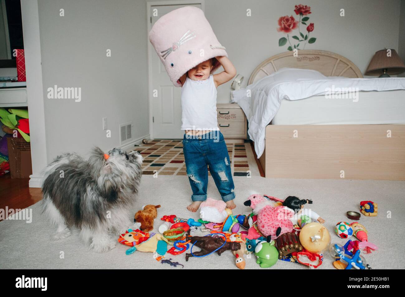 Cute adorable funny baby toddler girl with toy bin on head. Kid child playing with toys at home. Home authentic lifestyle. Kid with domestic animal Stock Photo