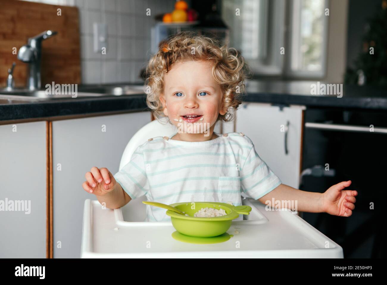 Cute adorable Caucasian curly kid boy sitting in chair eating cereal puree with spoon. Healthy eating for kids children. Toddler eating independently Stock Photo