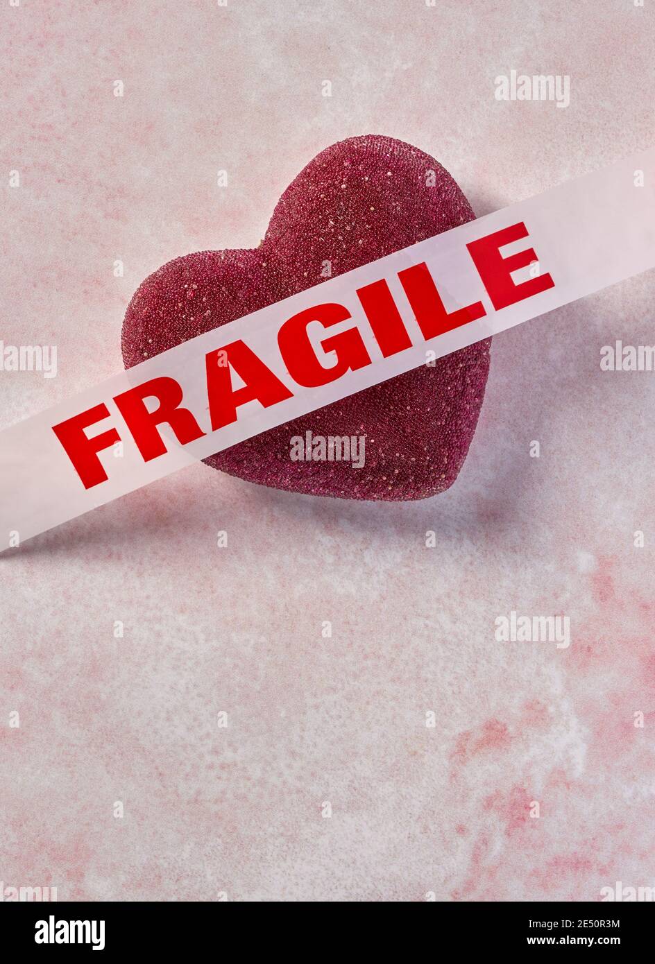 red heart with white tape with the word fragile in red ink across it sitting on a pink marbled background in vertical orientation Stock Photo