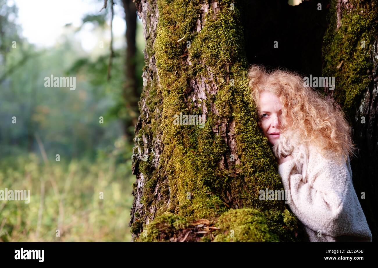 Portrait of a beautiful mature redhead woman in her forties with wild red curls and a fur scarf collar stands in an old willow tree, partially covered Stock Photo