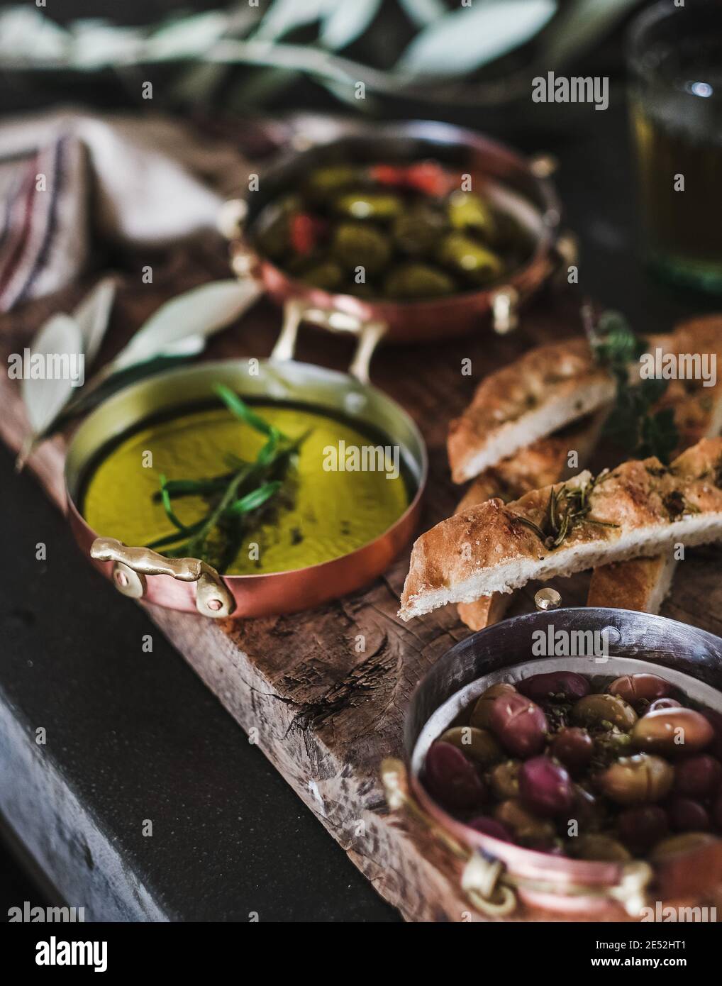 Pickled Greek olives, olive oil in copper jars and herbed focaccia slices on rustic wooden board, selective focus, close-up. Traditional Mediterranean meze appetizers platter Stock Photo
