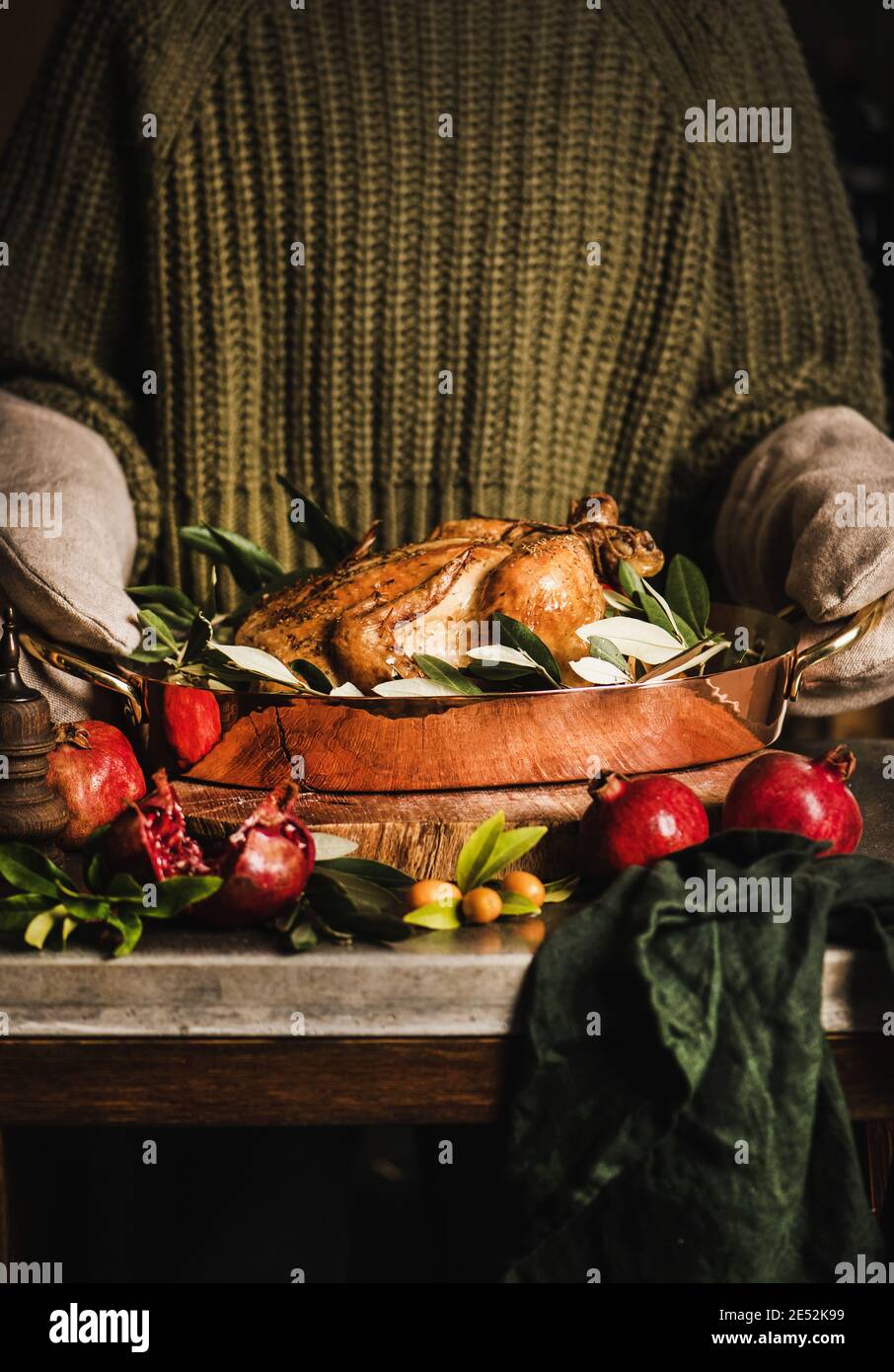 Woman in knitted sweater and kitchen mittens holding whole roasted chicken for holiday festive dinner in copper roasting tin with spices, herbs and fruit. Christmas or Thanksgiving Day cooking concept Stock Photo