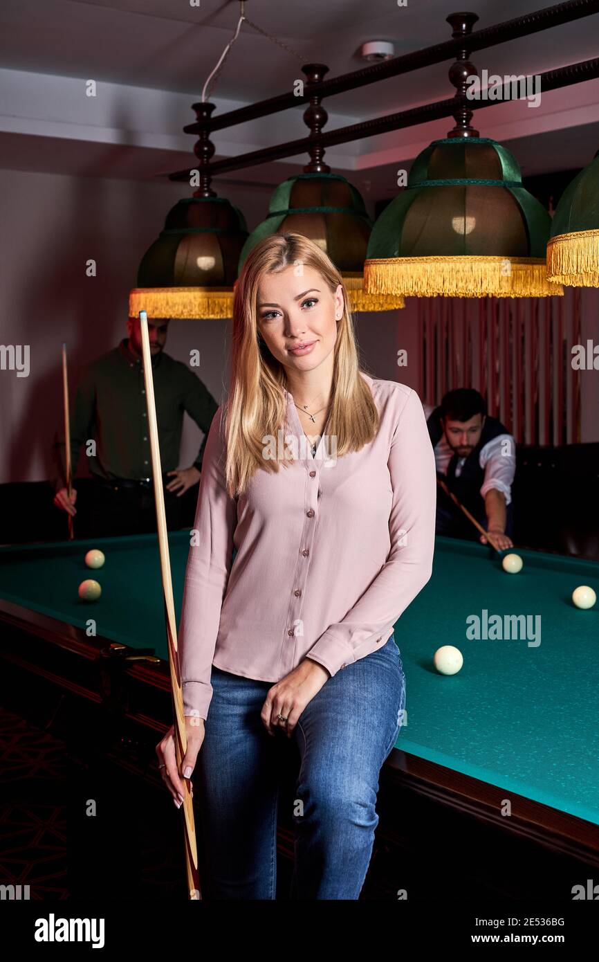 blond female came to spend pleasant time playing snooker, she is posing at camera, sitting on pool, billiards table Stock Photo