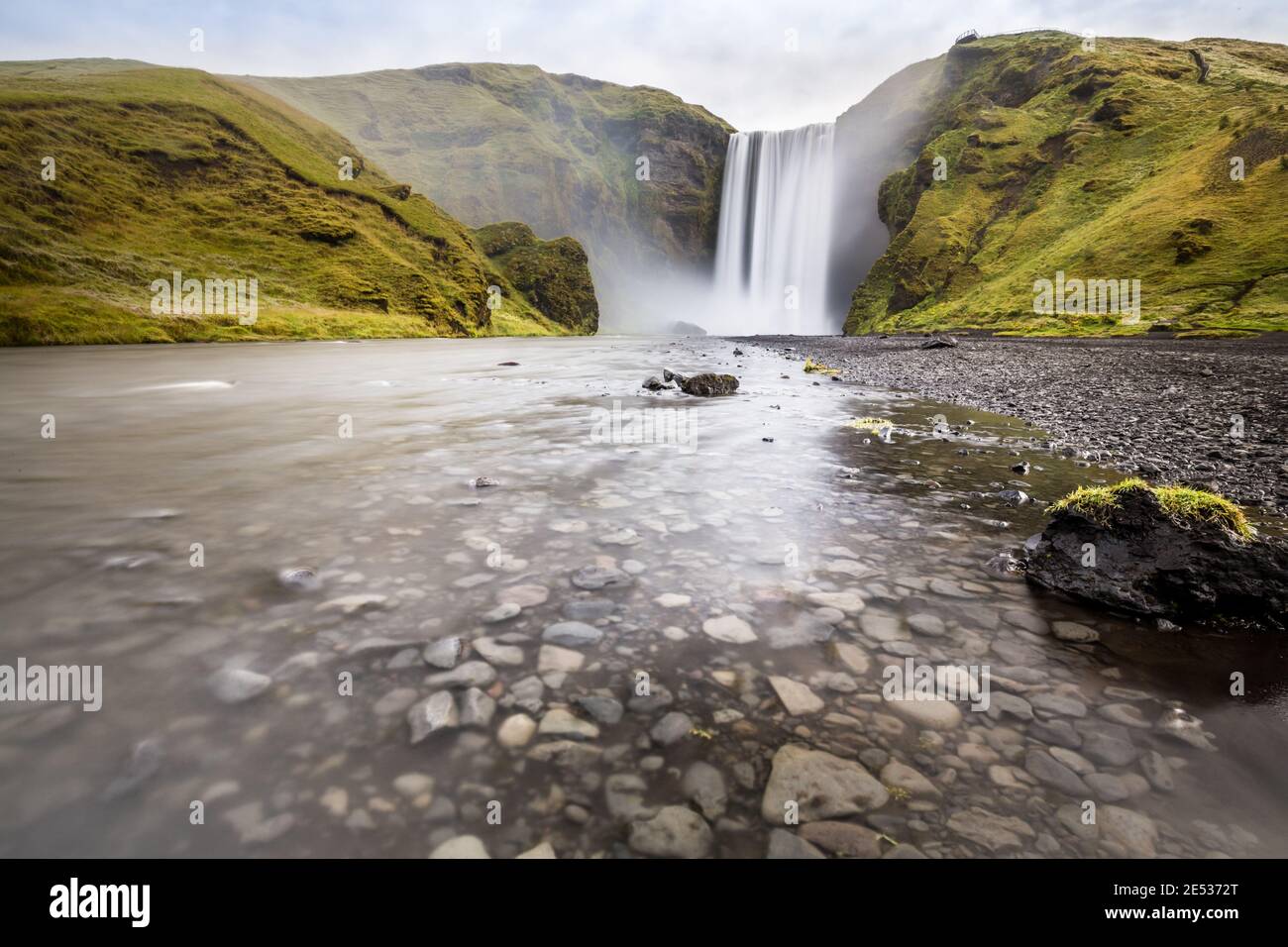 Iconic icelandic landscape with a stream flowing over pebbles in foreground and the Skogafoss waterfall, surrounded by green hills, in the background Stock Photo