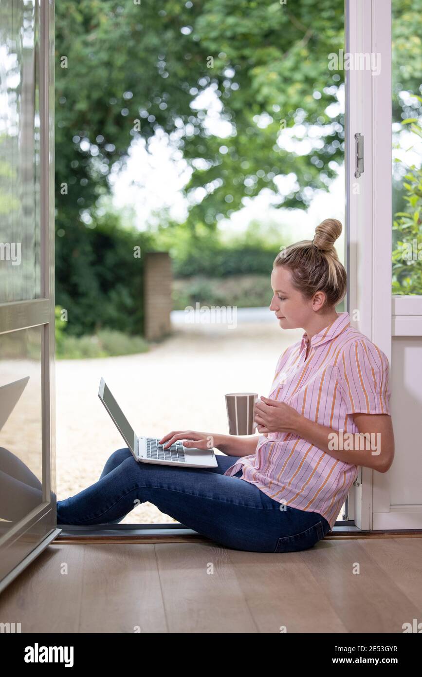 Businesswoman Working From Home On Laptop During Pandemic Lockdown Sitting In Open Front Door Stock Photo