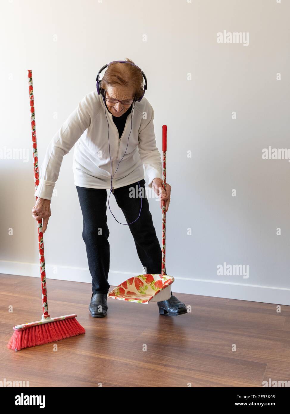 Cheerful 90 year old female enjoying music in headphones while sweeping floor with broom and doing housework Stock Photo