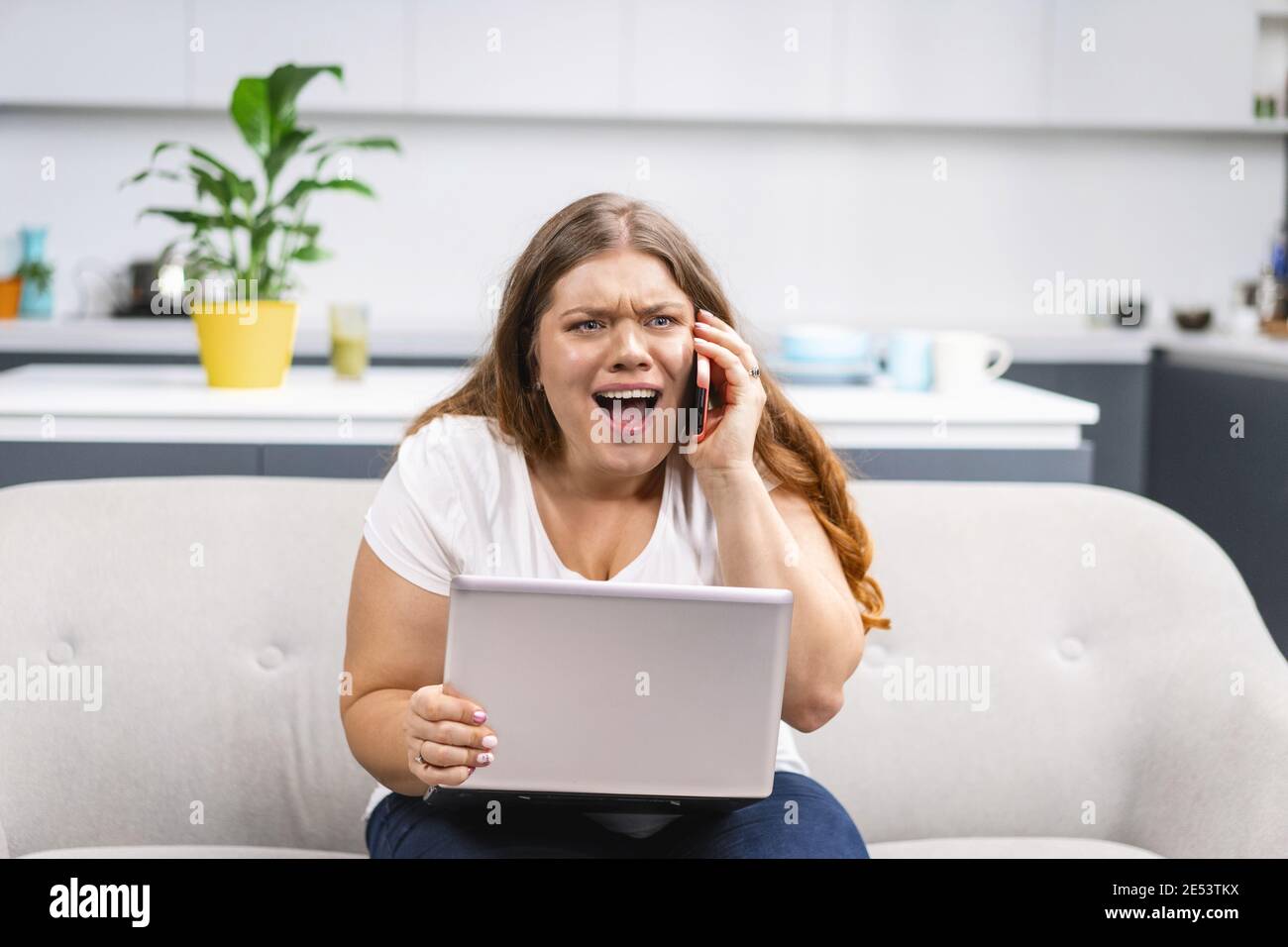Mad screaming talking on the phone young fat girl working on laptop sitting on the sofa at home with the kitchen on background. Self acceptance Stock Photo