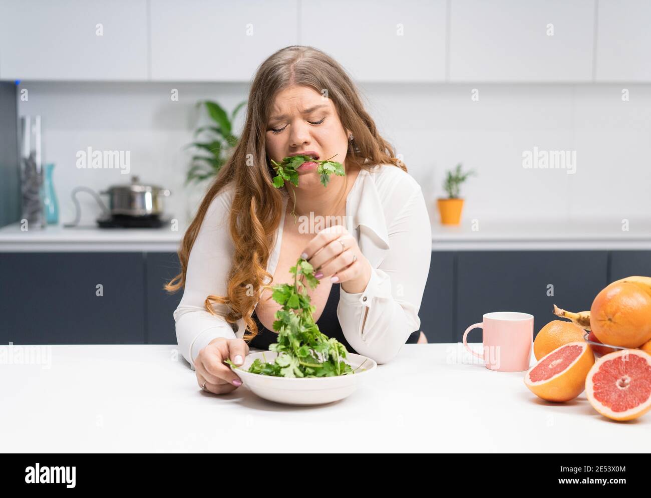 Young girl with weight problems holding fresh salad casting trying to chew  it. Curvy body young woman with long blond hair sitting on modern kitchen  Stock Photo - Alamy