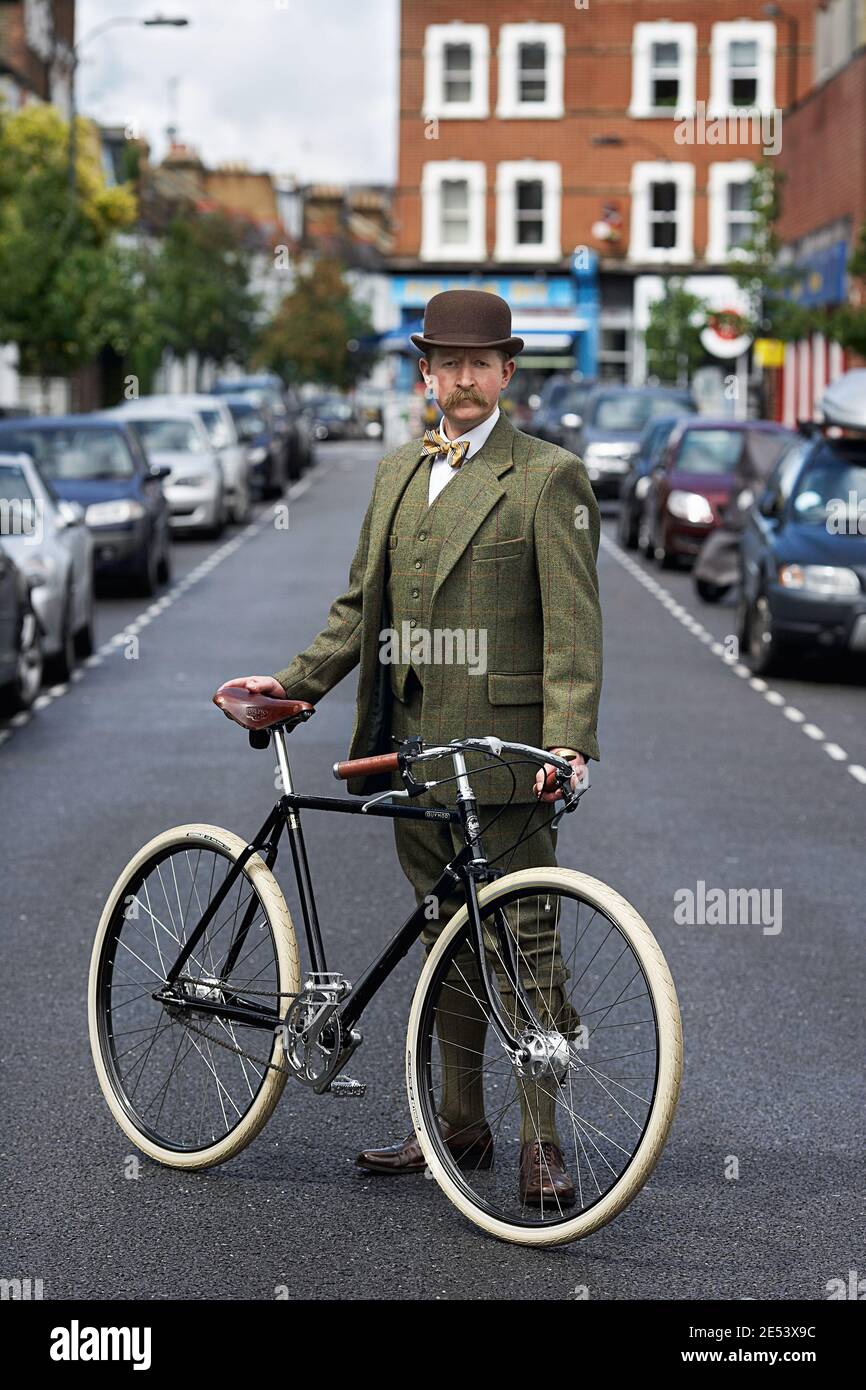Small street in London a elegant man standing beside Pashley Guvnor bike wearing suit with bowler hat and bow tie . Stock Photo