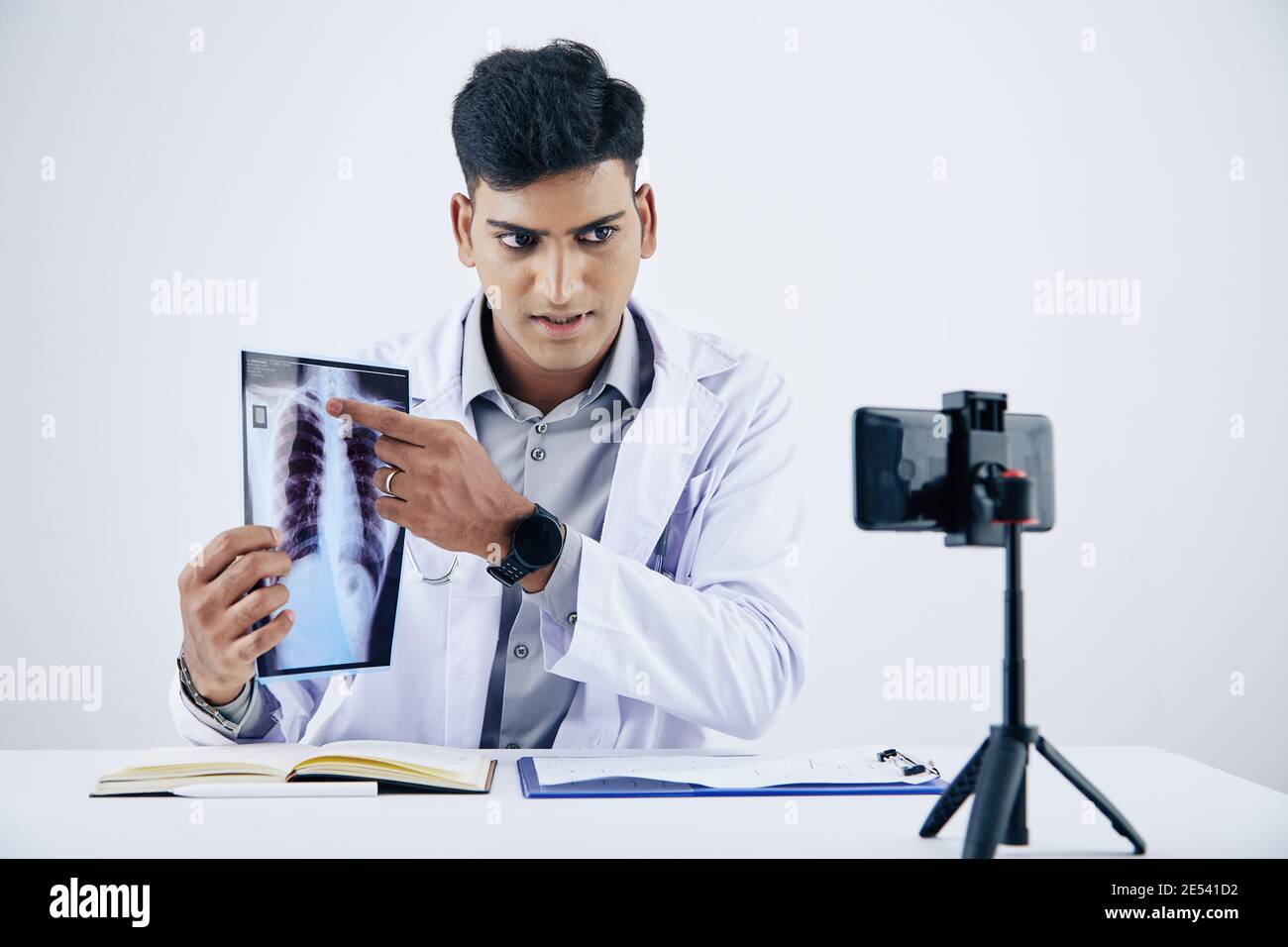 Serious young Indian doctor sitting at desk, pointing at chest x-ray when giving consultation to patient Stock Photo