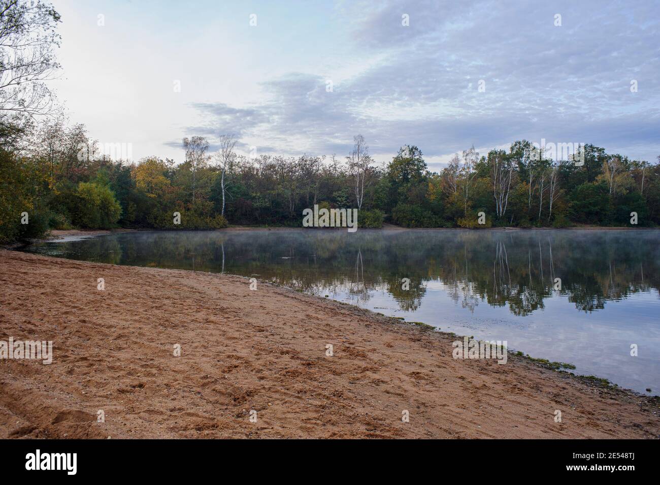 The beach of the amusement park is deserted. The trees on the shore are reflected in the calm water. A light mist rises in the cold morning air. Stock Photo