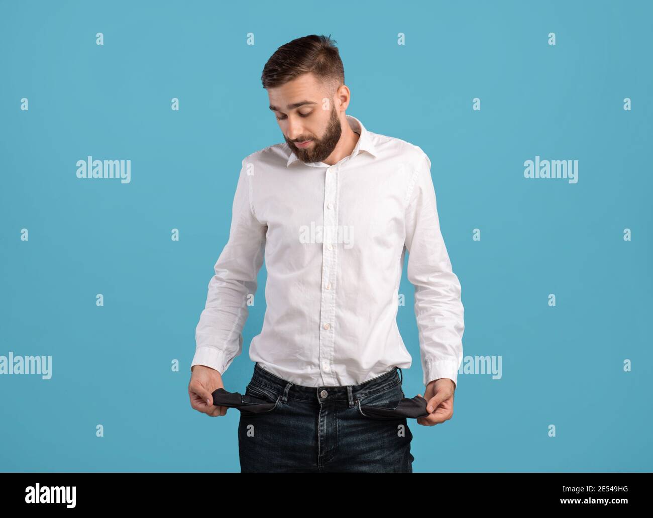 Young bearded man showing empty pockets on blue studio background. Economic depression concept Stock Photo
