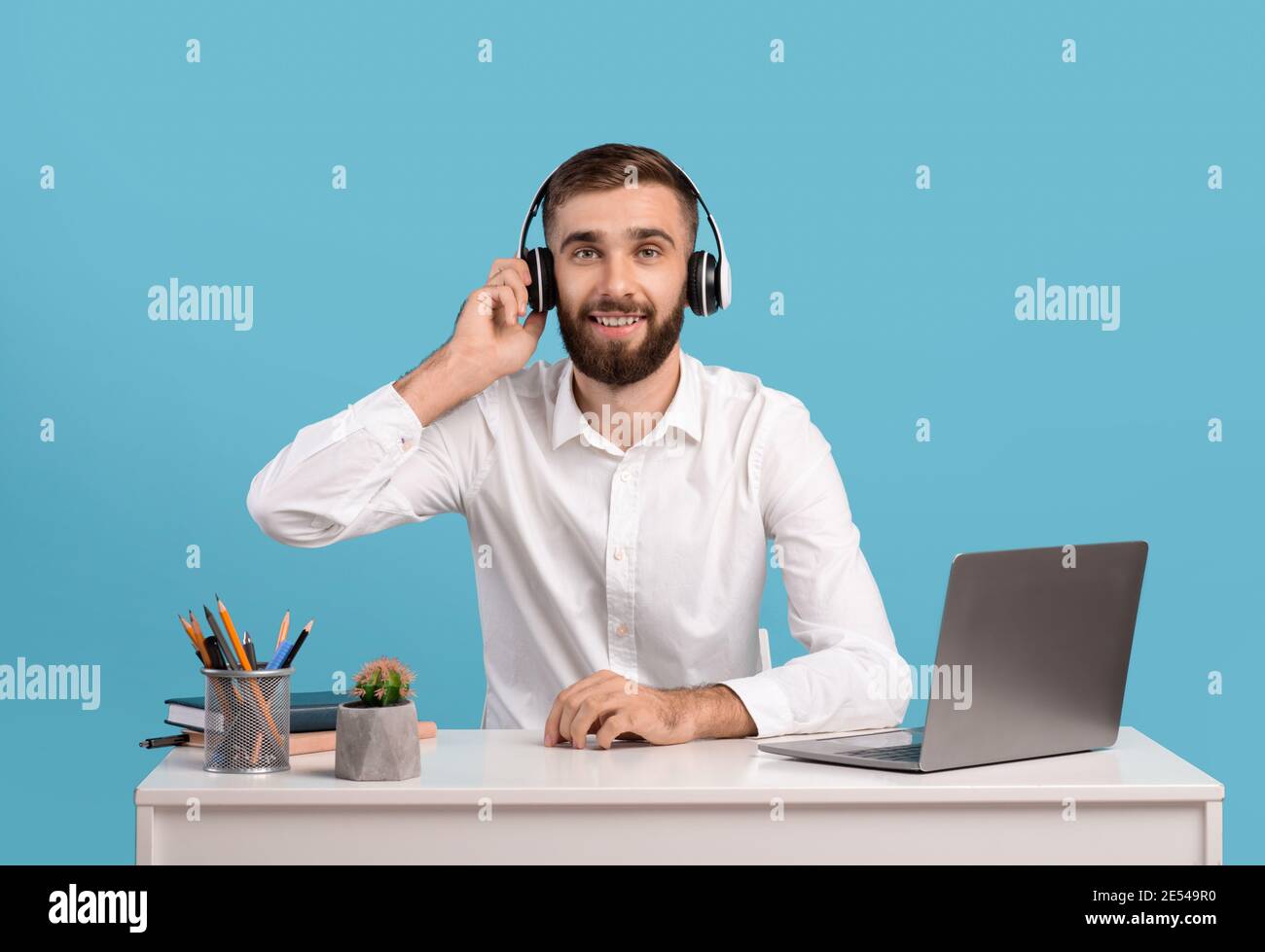 Bearded male helpline operator with headphones and laptop answering customer's call at desk over blue studio background Stock Photo