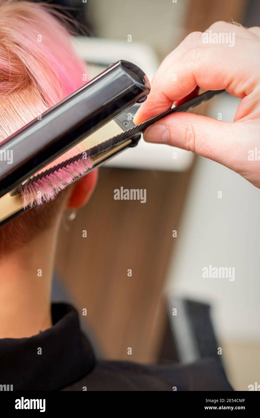 Close up of the hairdresser straightening the short pink hair of a female client with a hair straightening iron in a beauty salon Stock Photo