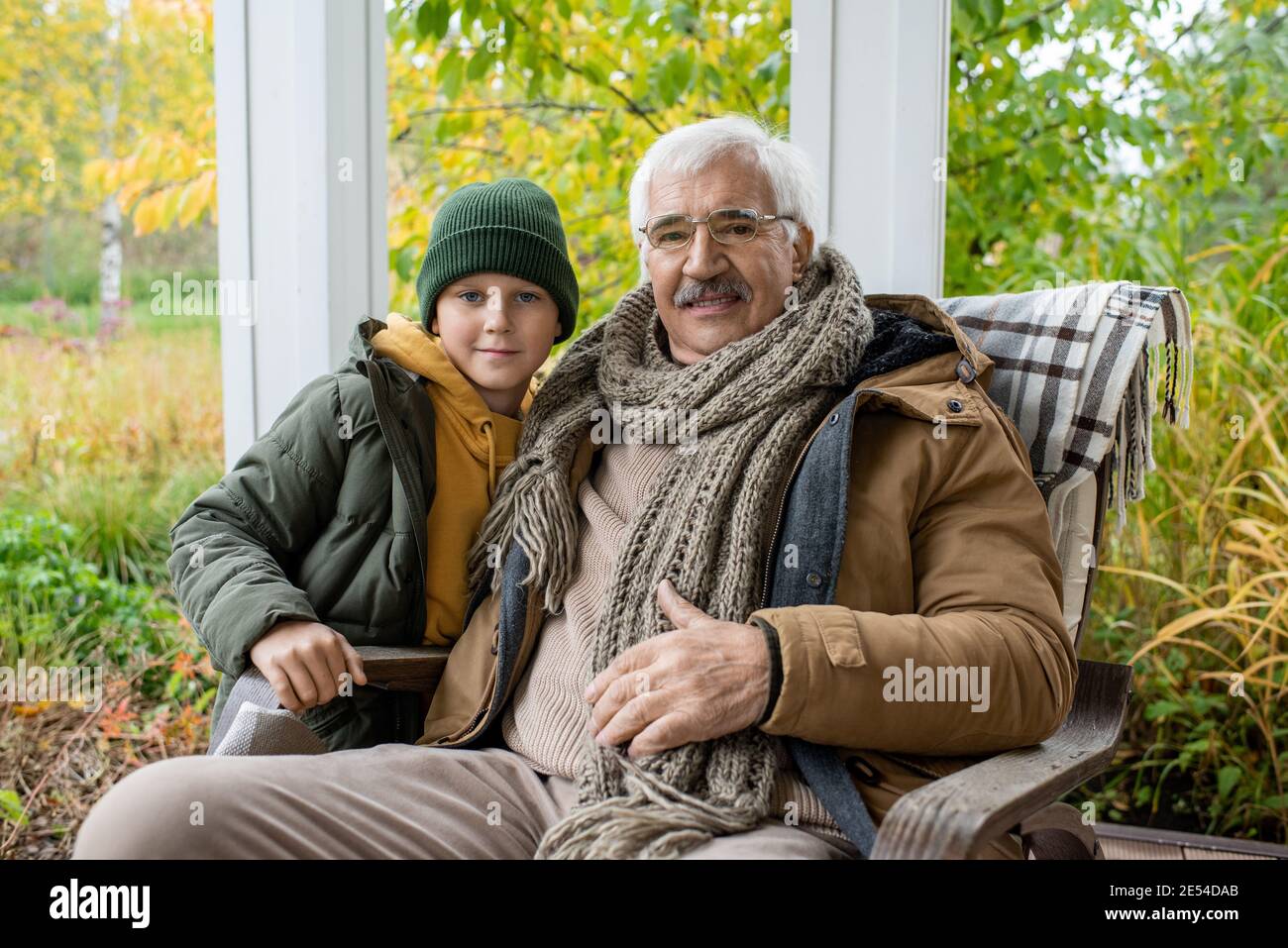 Cute schoolboy in beanie and warm jacket and his grandfather in rocking  chair looking at you while relaxing against natural environment Stock Photo  - Alamy