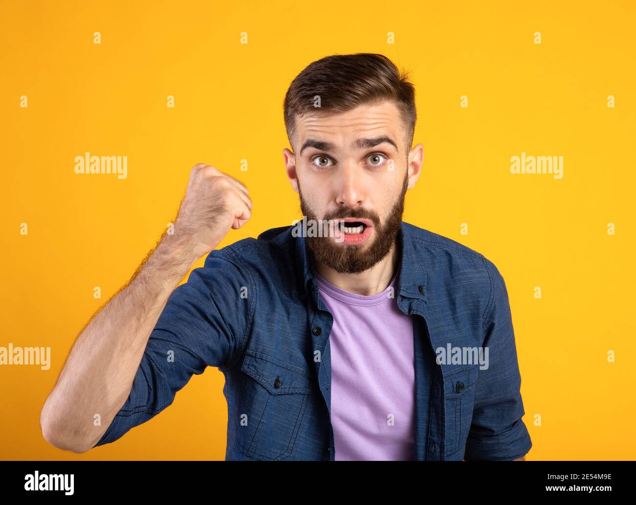 Portrait of young angry guy shaking clenched fist at camera and shouting, showing his protest over orange background Stock Photo
