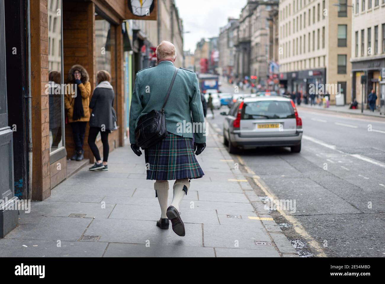 Edinburgh Scotland - October 2019 Scottish man walking along high street in tartan kilt with bag and green jacket through city centre from behind look Stock Photo