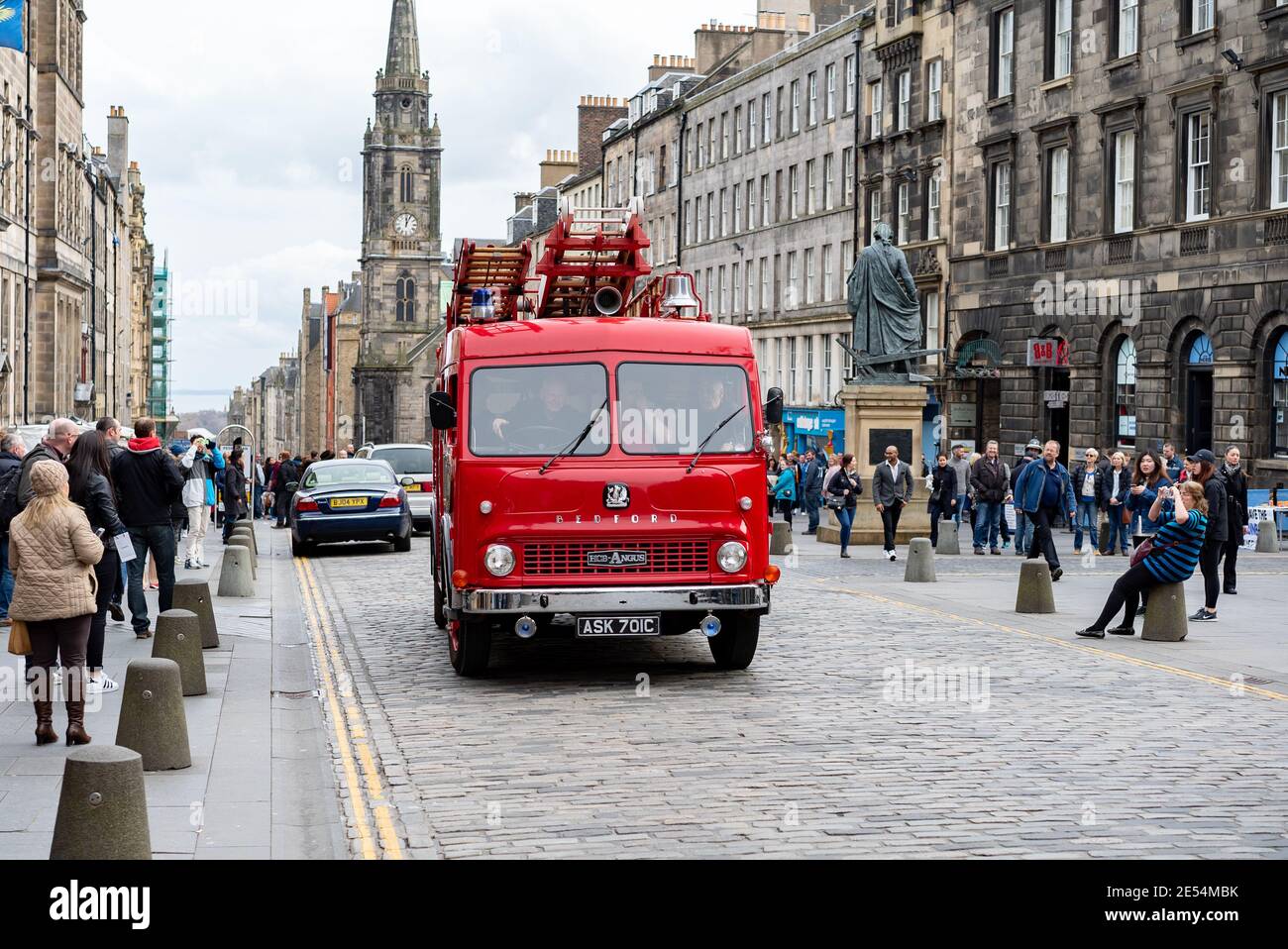 Classic old red silver Bedford fire engine service with wooden extendable ladder driving on high street cobble stones shoppers watching church sunny Stock Photo