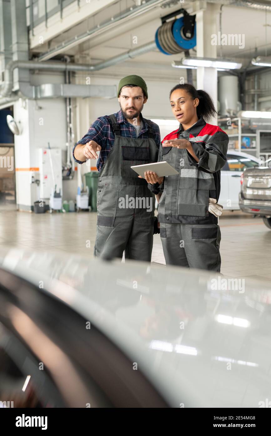 Young repairman and his female colleague in uniform discussing characteristics of car in workshop while using data from digital tablet Stock Photo
